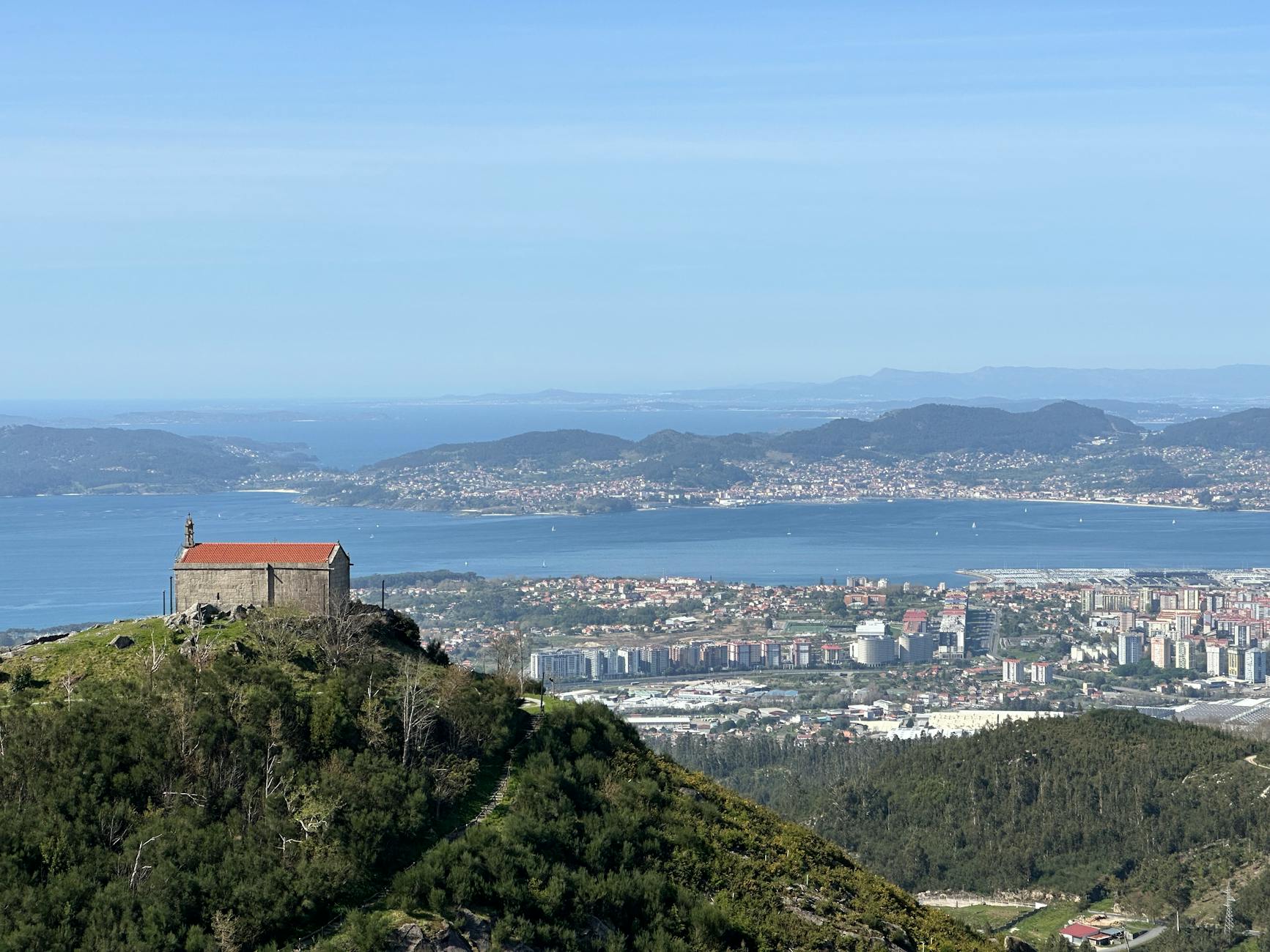 Aerial view of Vigo city, bay, and lush hills on a clear summer day, including a solitary building atop a hill.