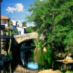 Beautiful stone bridge in Allariz, Spain, reflecting in a tranquil river surrounded by lush greenery and historic architecture.