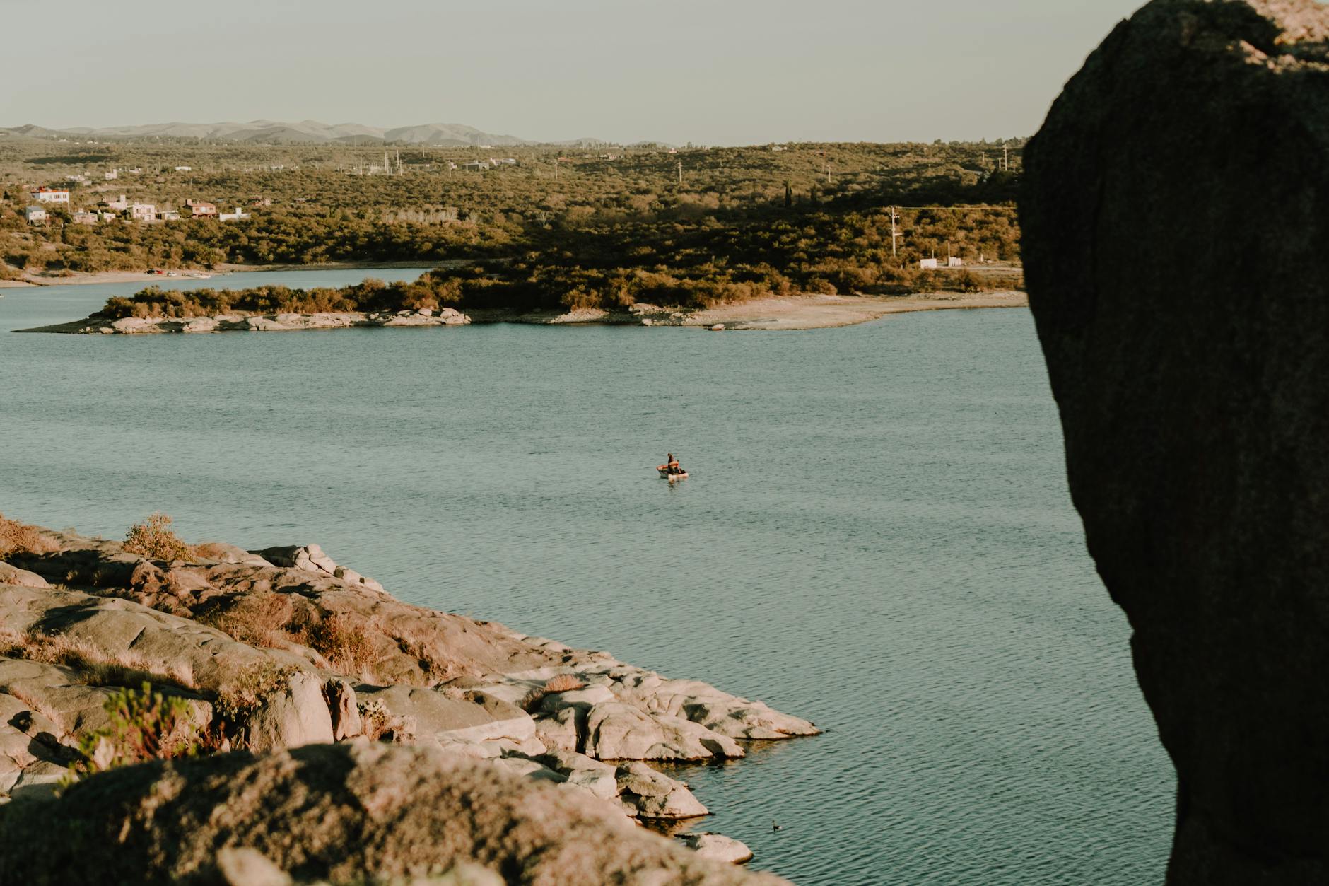 Serene view of a lone kayaker paddling on a lake near Capilla del Monte, surrounded by rocky landscape.