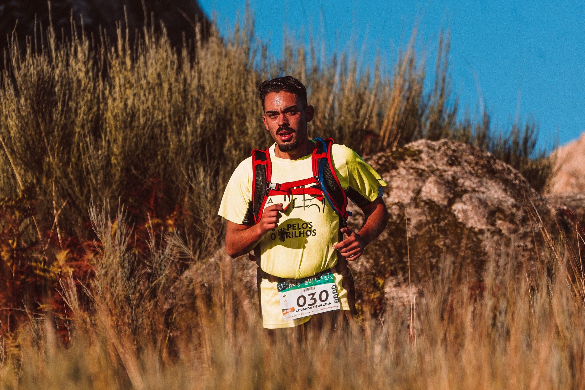A man participates in a trail marathon, running through rugged terrain in bright sunlight.