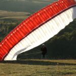 A paraglider with a red and white canopy prepares to take off from a grassy hill in a hilly landscape.