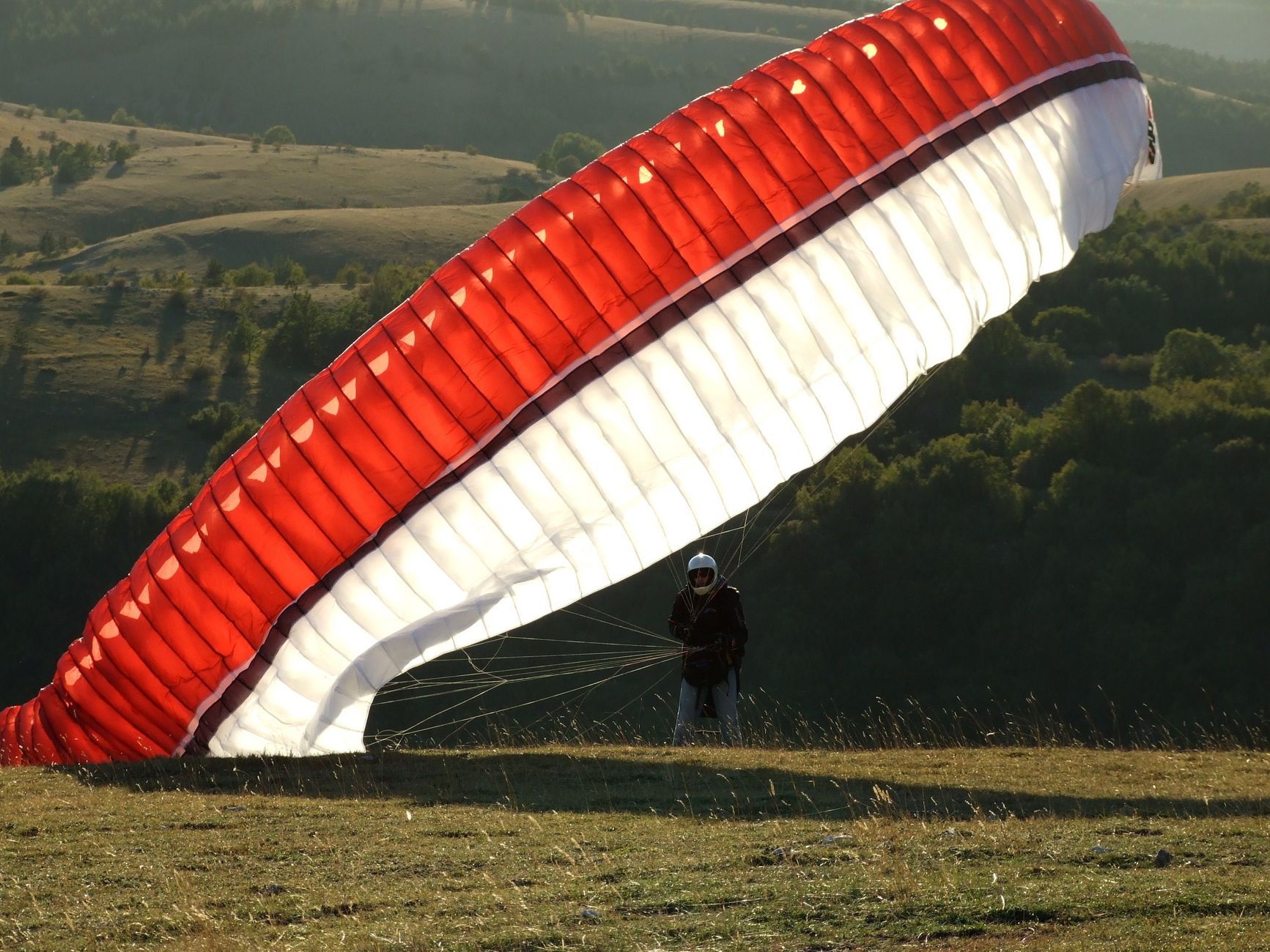 A paraglider with a red and white canopy prepares to take off from a grassy hill in a hilly landscape.