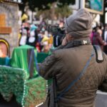 A man in a beanie and puffer jacket photographing a lively outdoor event.