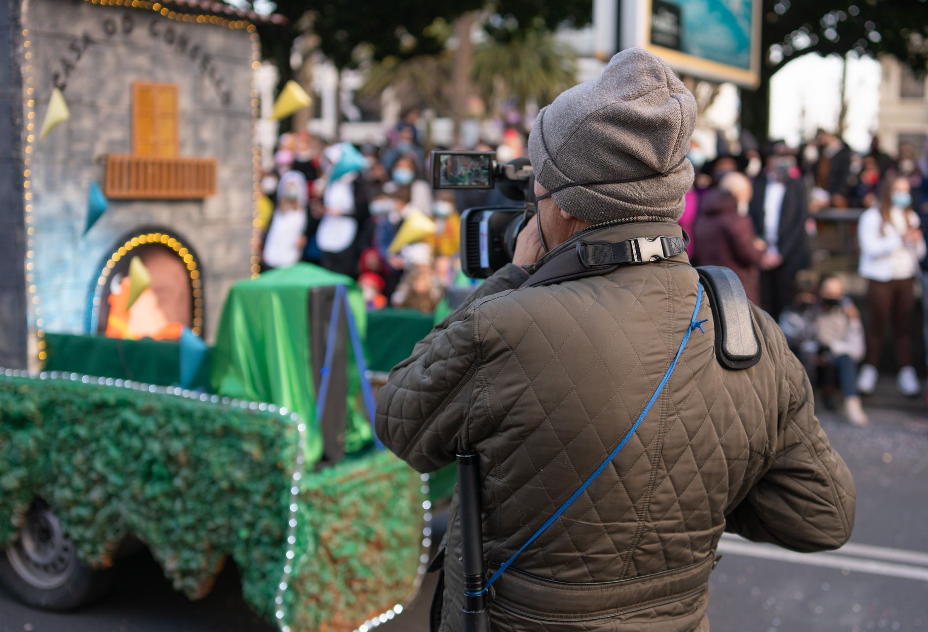 A man in a beanie and puffer jacket photographing a lively outdoor event.