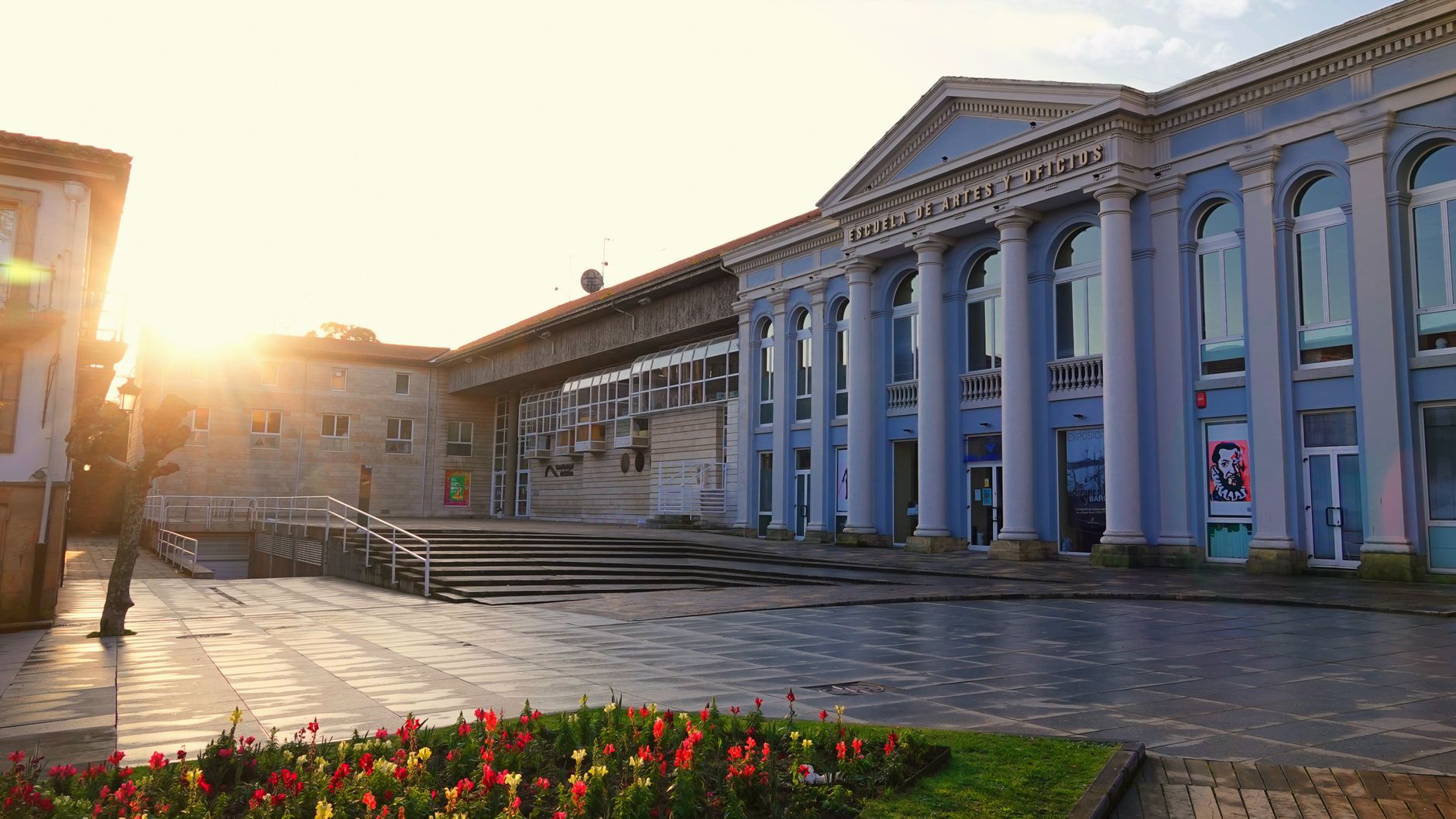Sunny early morning view of a classical building with columns in an urban environment, capturing vibrant flowers and modern architecture.