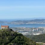 Aerial view of Vigo city, bay, and lush hills on a clear summer day, including a solitary building atop a hill.