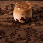 A cold iced coffee with chocolate syrup surrounded by coffee beans on a rustic wooden table.