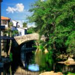 Beautiful stone bridge in Allariz, Spain, reflecting in a tranquil river surrounded by lush greenery and historic architecture.